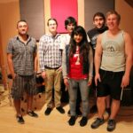 a group photo of the band among the brave along with john garcia and hector gomez in the tracking room of let down studio with a partially set up drum set in the background.