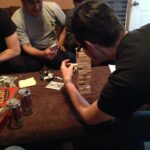 a photo of the back of john garcia playing jenga with beer cans on a table and a pizza with christopher olivas across from him.