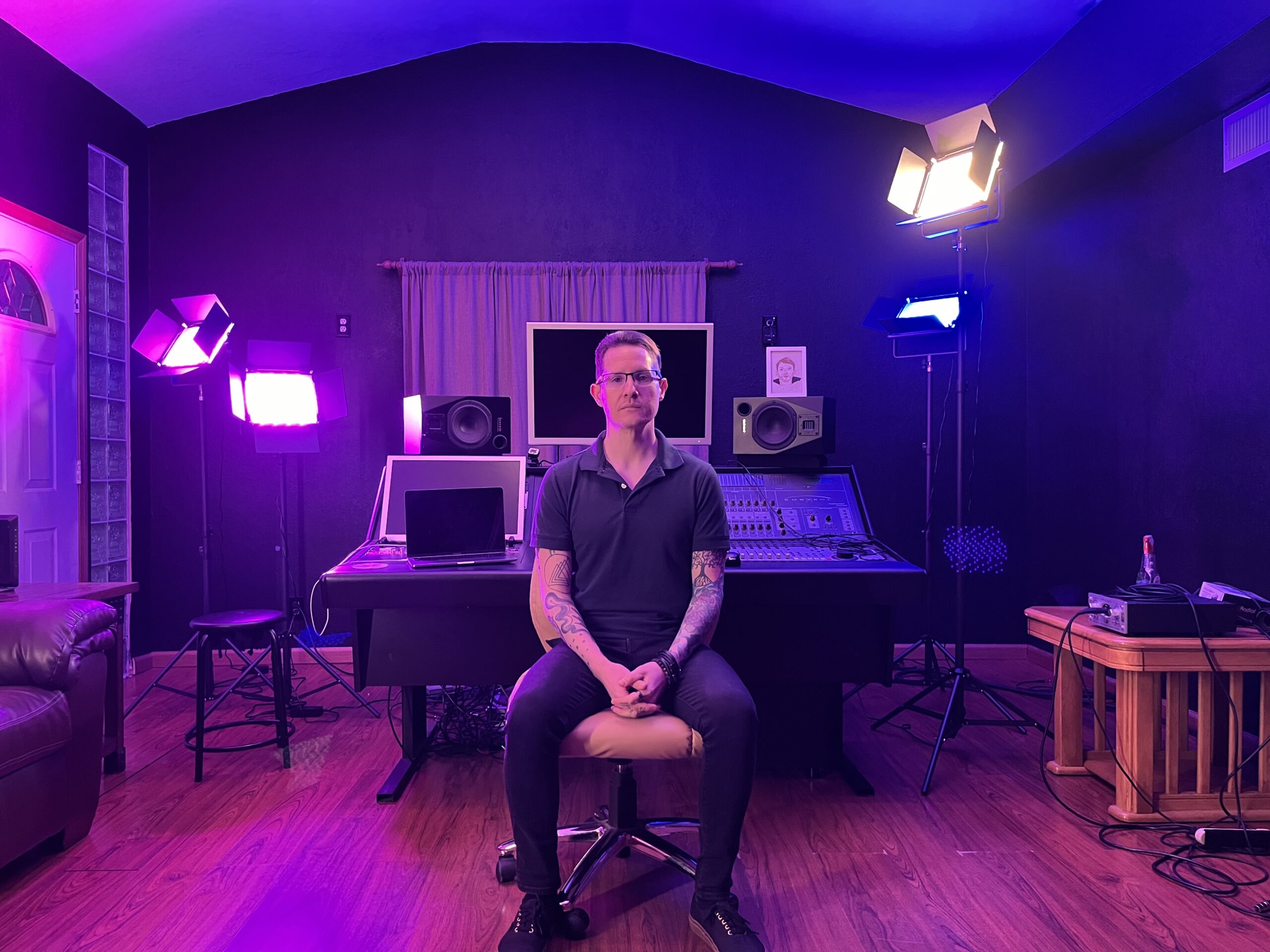 a front view of John Garcia sitting in front of the mixing board at let down studio with 4 blue and purple lights illuminating the room.