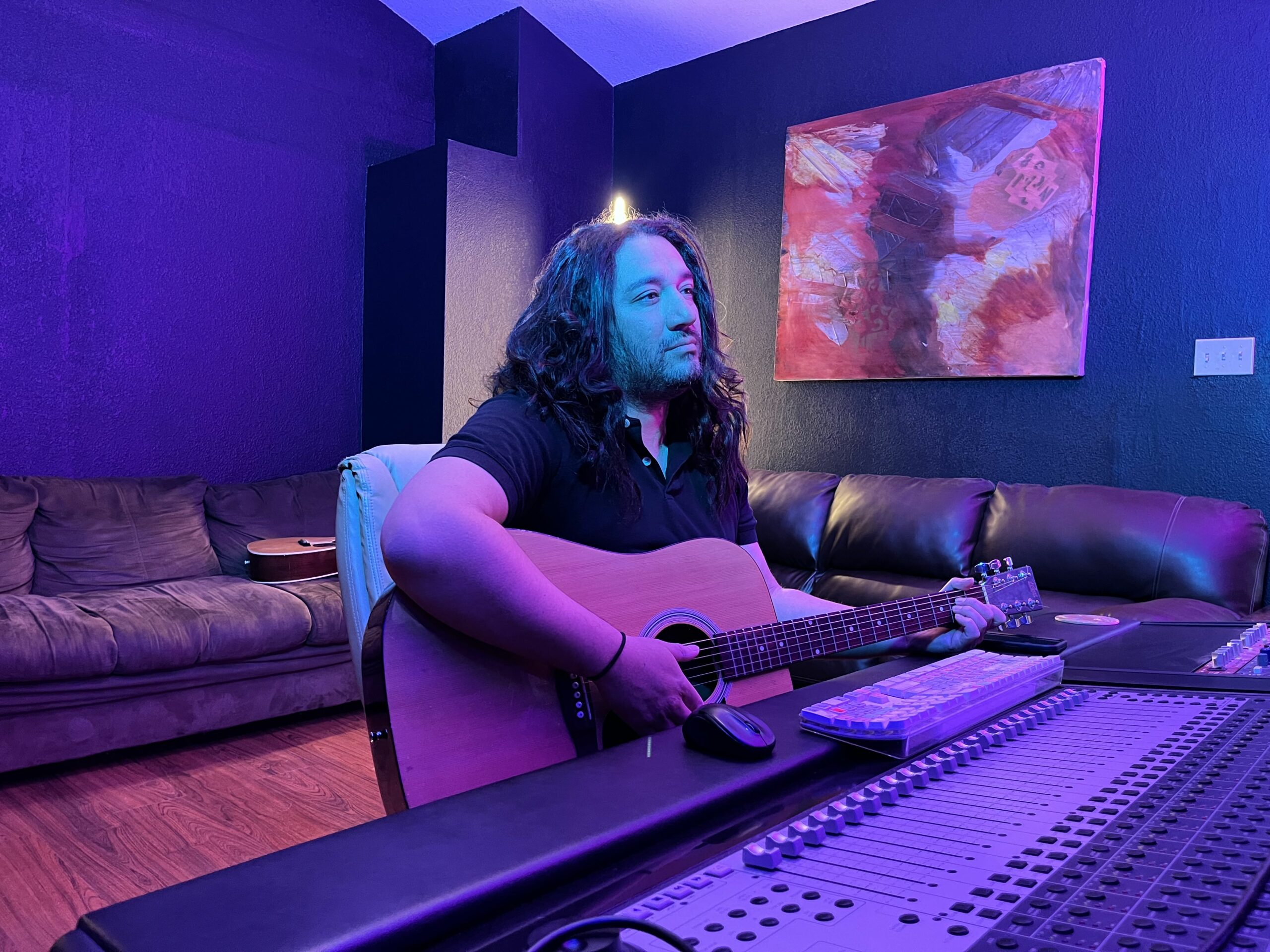 a photo of hector gomez sitting in front of the mixing board with an acoustic guitar with a blue light illuminating the room.