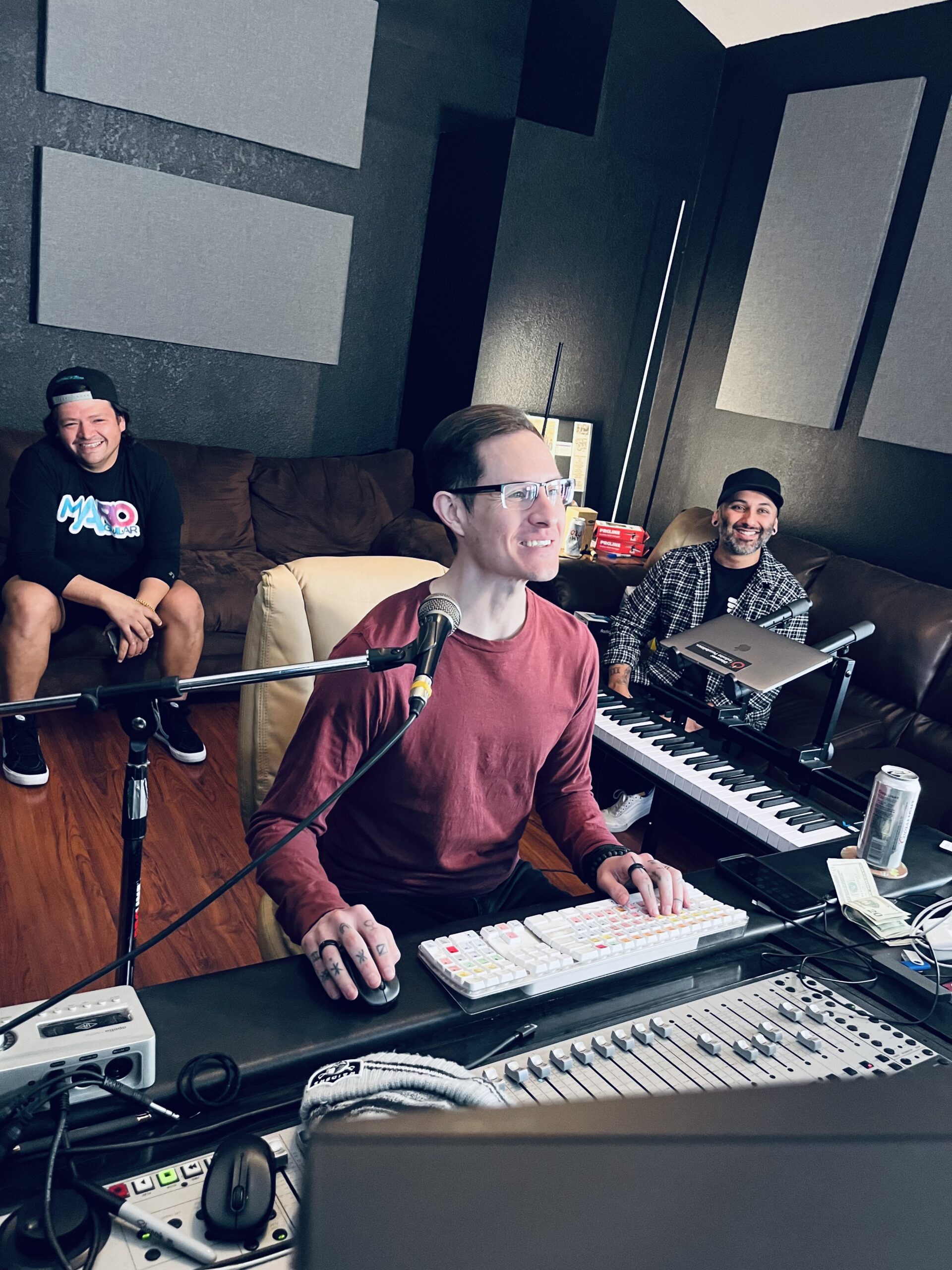 A front view of John Garcia recording on a mixing board, Miguel Alcala sitting and smiling on a couch, and Alex Hernandez sitting and watching the recording on a brown couch.